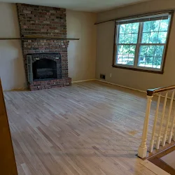Empty living room with hardwood flooring, brick fireplace, and large window